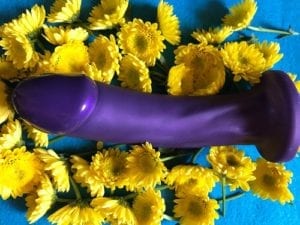Tantus Adam Super Soft, purple, lying sideways in yellow flowers