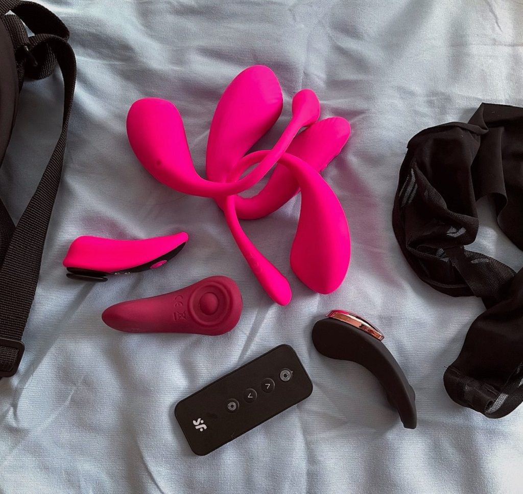 Wearable Bluetooth vibrators on bed featuring Lovense Lush 2, Lovense Lush 3, Lovense Quake, Lovense Ferri, Satisfyer Sexy Secret, and Satisfyer Little Secret public play sex toys