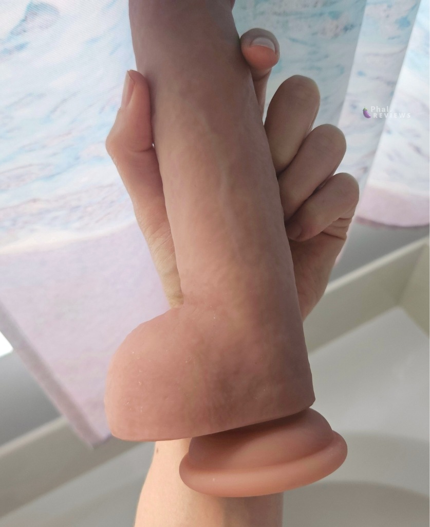 Photo of tan Solina Smootha dildo's silicone lower shaft showing smooth "balls" and suction cup, held in reviewer's hand in front of ocean print bathroom curtain