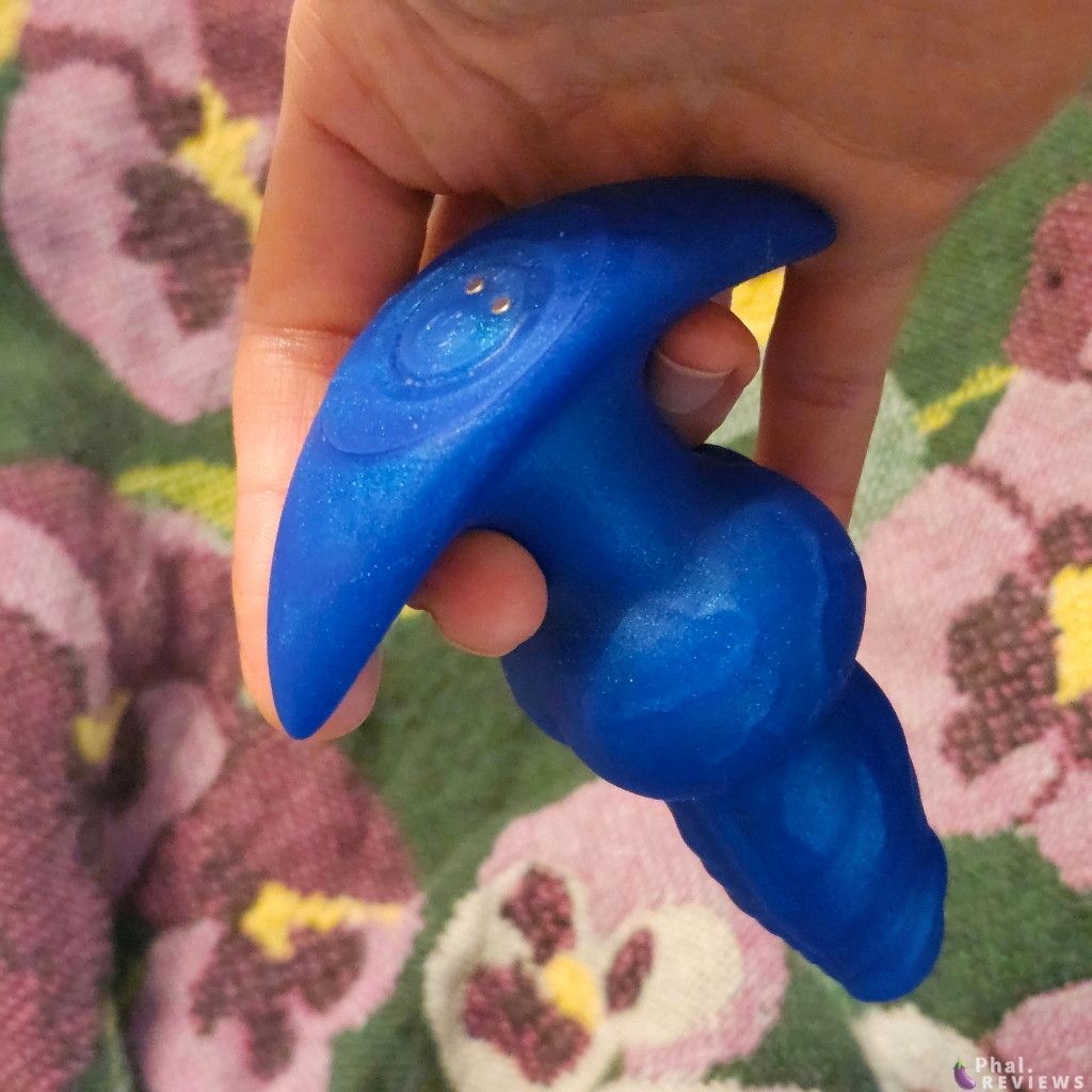 Closeup on flared shape of blue silicone plug in reviewer's hand, showing base plus power button and charging contacts, held over floral throw blanket