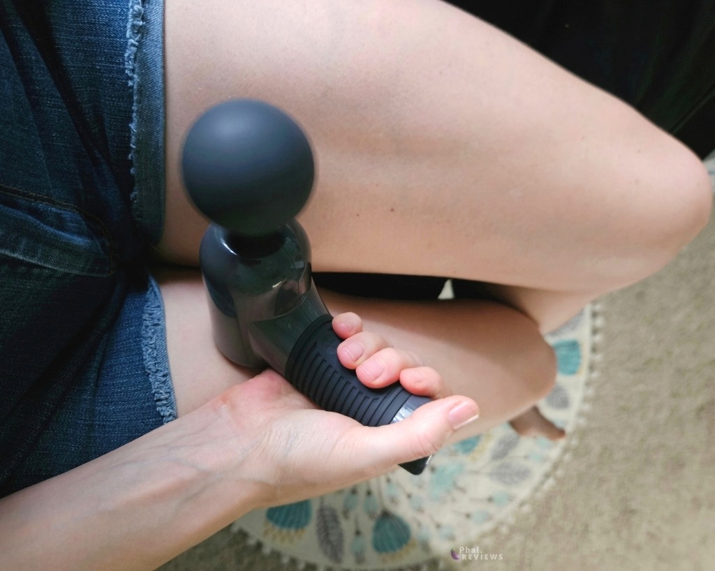 Large Massage Gun's round silicone blurred as it vibrates, held upward by reviewer's hand and forearm over her denim shorts