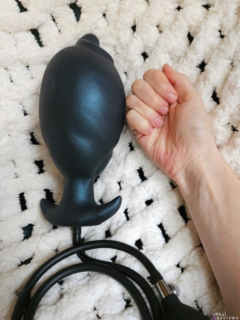 Fully inflated black plug with blue-green shimmer next to reviewer's clenched fist, over white crocheted blanket. Black coiled tubing is attached underneath the silicone plug.