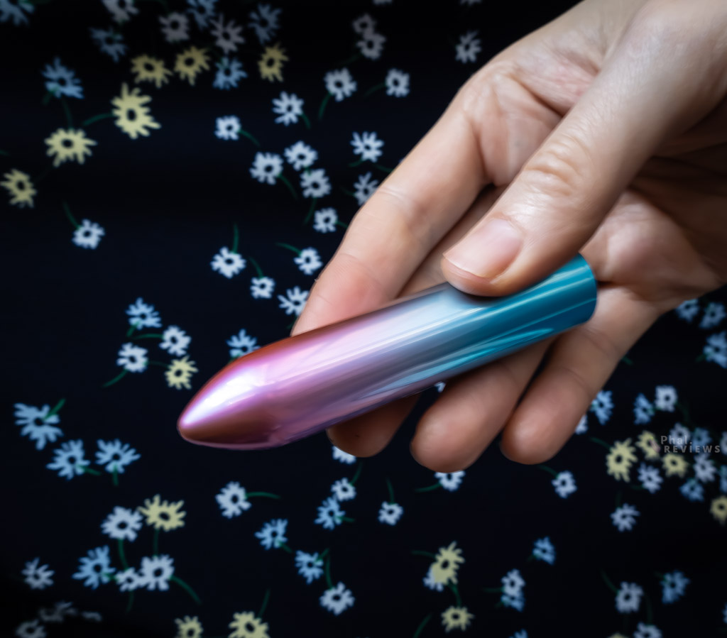 Light hitting pink-to-blue gradient colors of Shimmr vibrator held diagonally in reviewer's fingers over dark blue background wtih white and yellow flower design