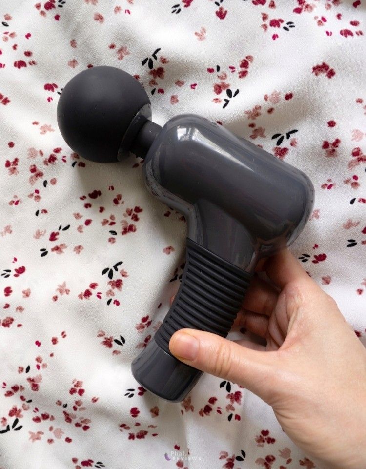 Black silicone headed plus gray body massager with its handle being reached for by a hand, as it rests over an abstract floral shirt with crimson and pink stylized designs